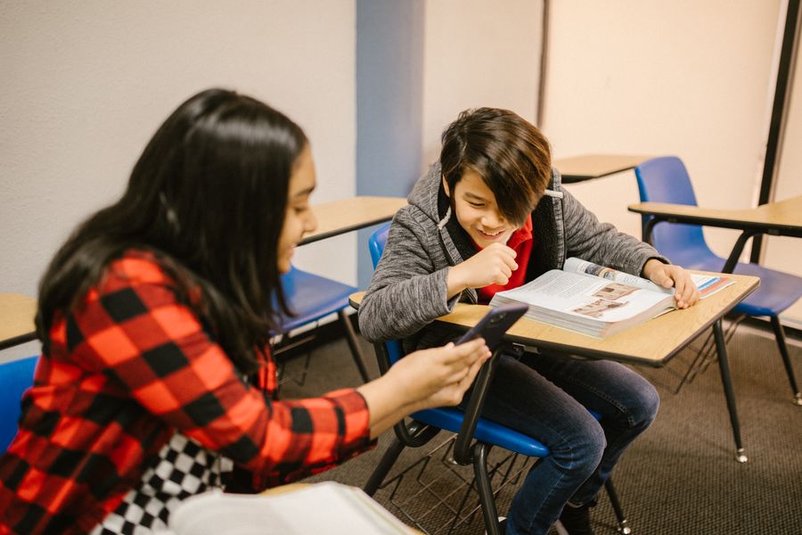 Two students interact with a smartphone in a school classroom setting.