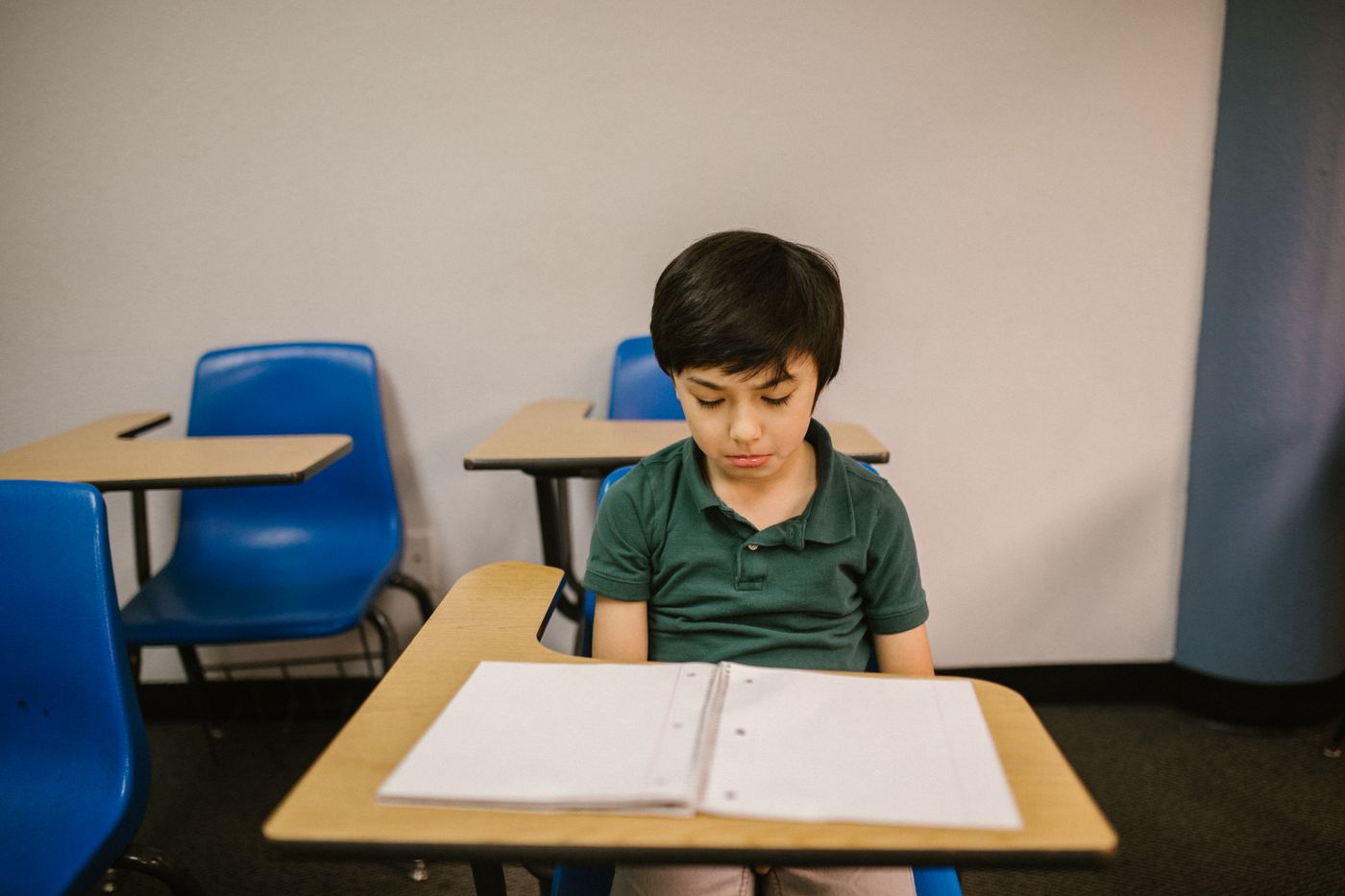 A young boy sitting alone at a classroom desk, looking down and withdrawn.