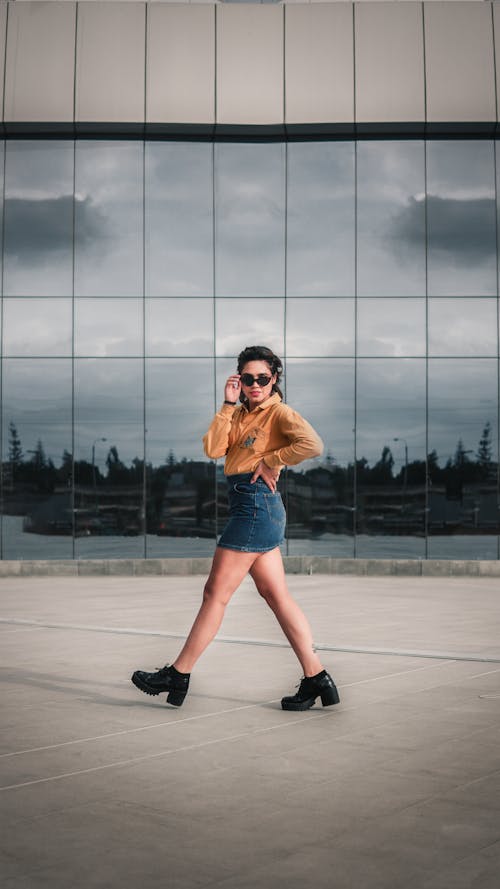 Free Woman in Brown Long Sleeve Shirt and Blue Denim Skirt Walking on the Street Near Glass Building Stock Photo