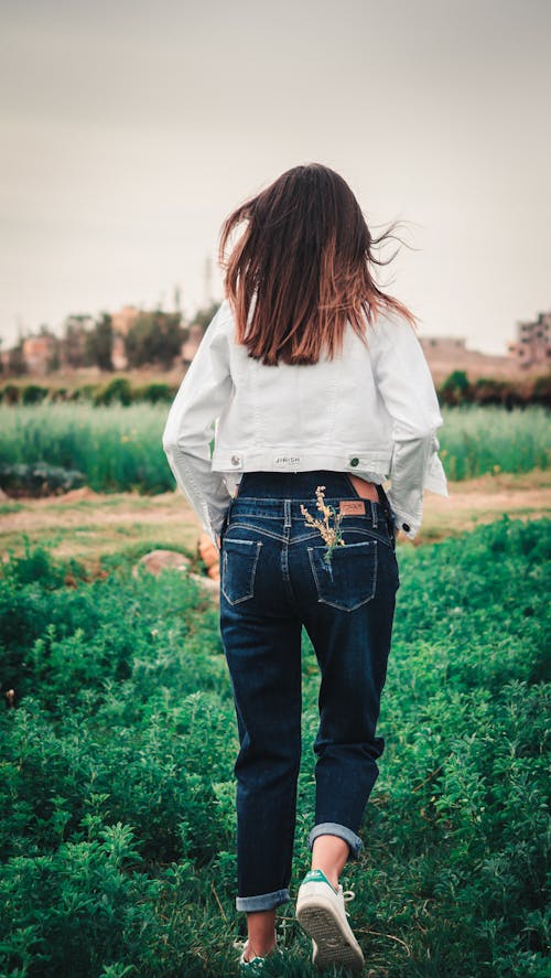 A Back View of a Woman in a White Jacket and Denim Pants Walking on a Field
