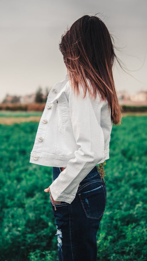 Free Woman in White Jacket and Blue Denim Jeans Standing on Green Grass Field Stock Photo