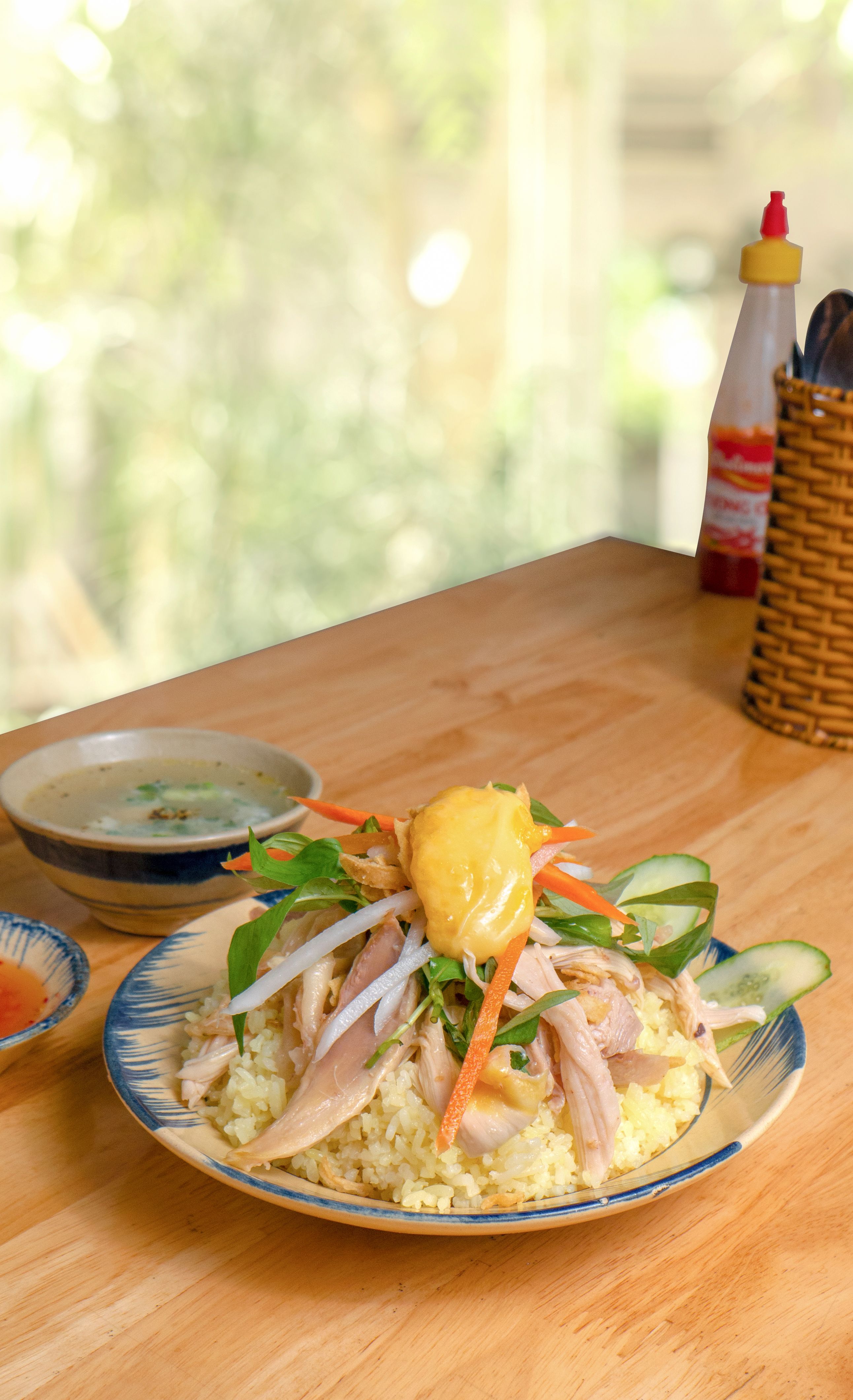 A white bowl of pale golden chicken and rice soup with soft diced carrots and shredded chicken, steam rising, set on a light wooden table.