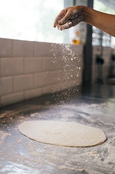 A hand sprinkles flour on pizza dough on a surface in a kitchen, capturing the art of cooking.