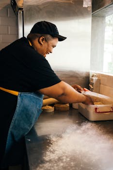 Smiling chef kneading dough in an industrial kitchen, wearing a black cap and apron.