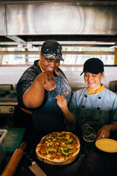 Two chefs joyfully prepare and sprinkle toppings on a delicious pizza in a bustling restaurant kitchen.