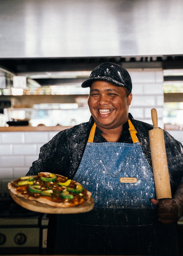 A Man In Blue Apron Holding A Rolling Pin And Pizza On Wood Tray