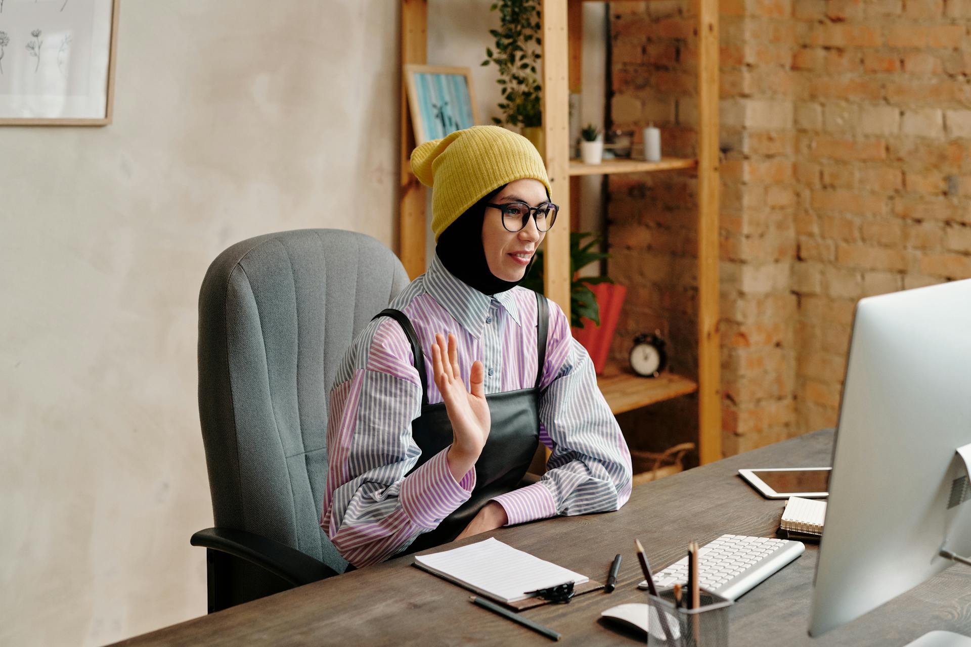 A woman wearing a yellow hat waves during a video call in a cozy home office setting.