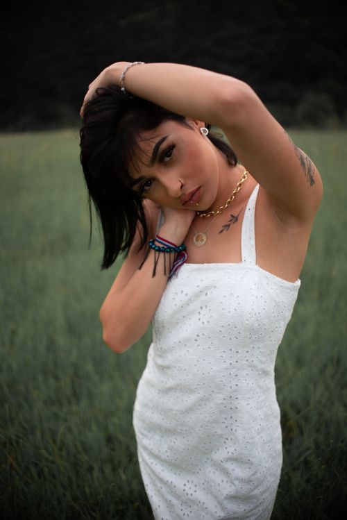 Free Charming tattooed female wearing white dress looking at camera while standing on grassy meadow with hand on head in nature Stock Photo