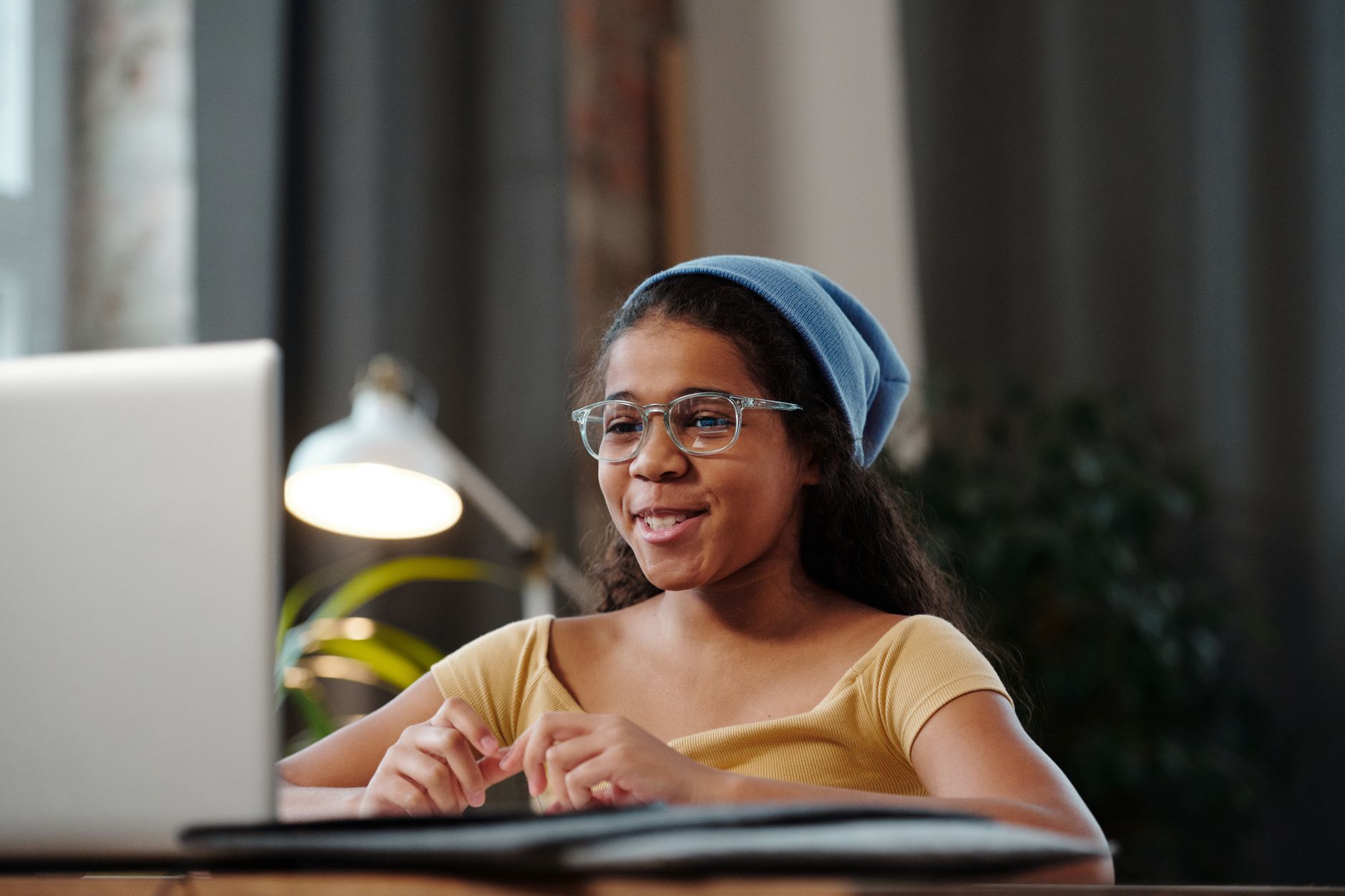 Jeune fille à lunettes et bonnet bleu souriant devant un ordinateur portable lors d'une discussion vidéo