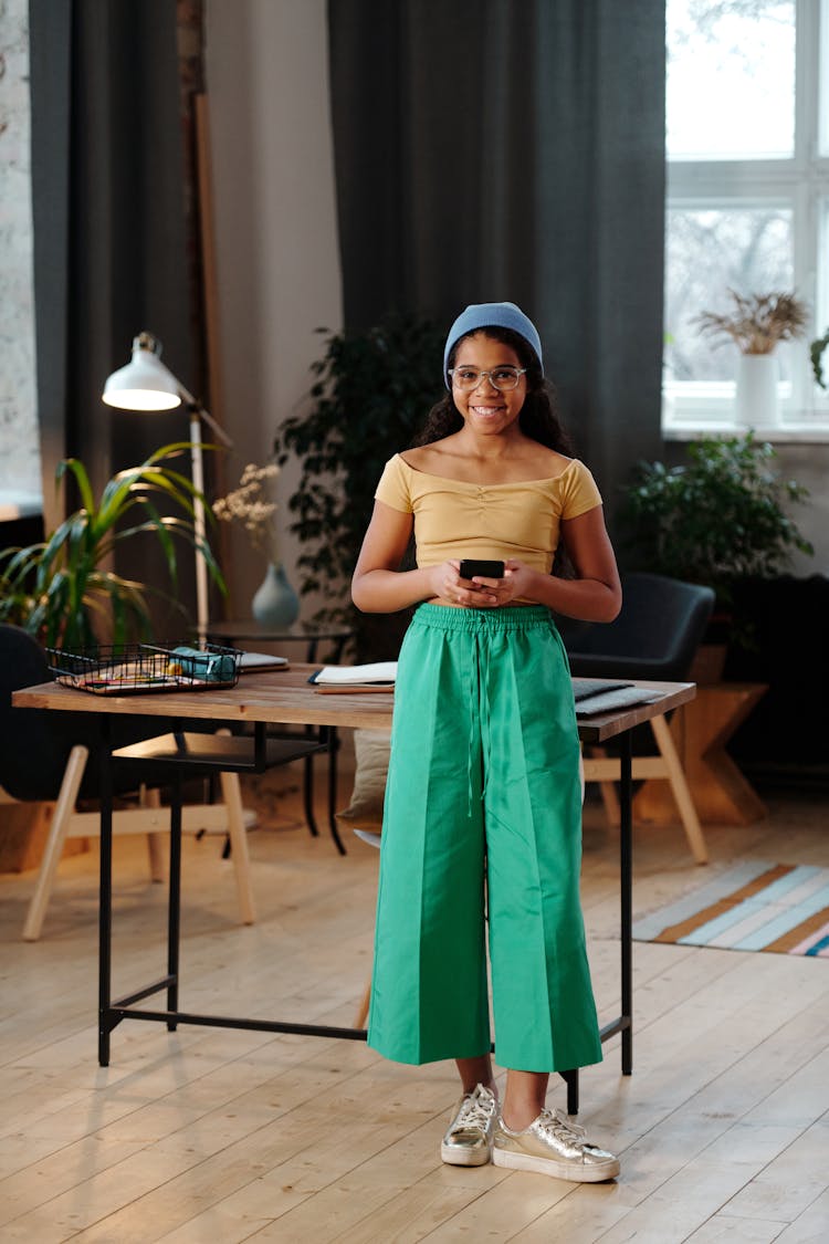 Girl Smiling And Holding A Phone While Standing In Front Of A Desk At Home 