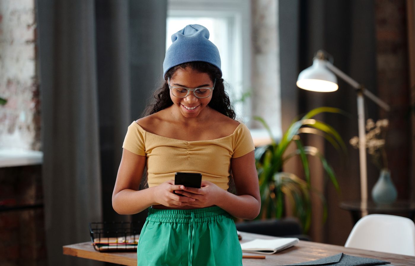 Cheerful teenage girl with eyeglasses using smartphone indoors, smiling and enjoying a digital moment.