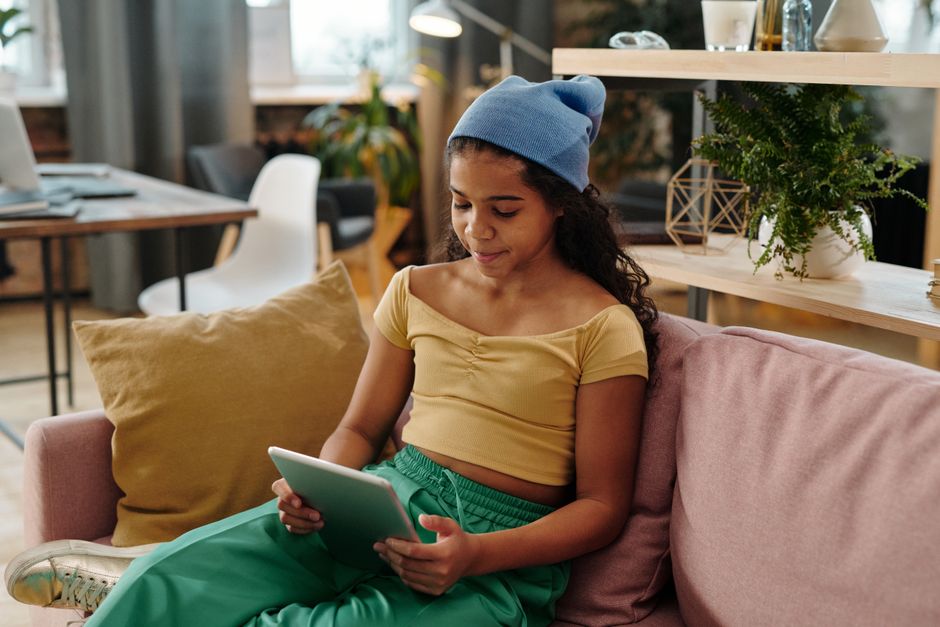 A young woman in casual attire sits on a sofa, using a digital tablet in a cozy living room.