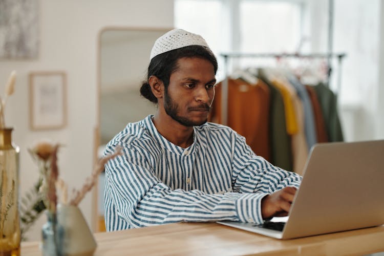 A Bearded Man In Kufi Cap Using His Laptop