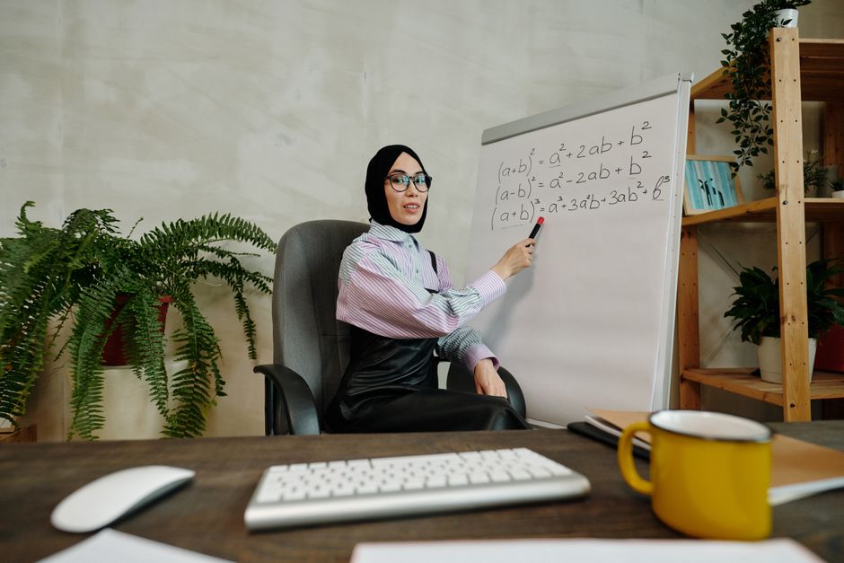 Female teacher explaining algebraic formulas on a whiteboard in a home office setting.