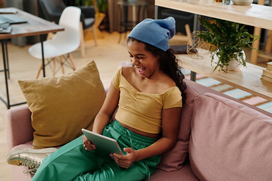 Happy teenager wearing beanie, sitting on a sofa, and enjoying a tablet indoors.