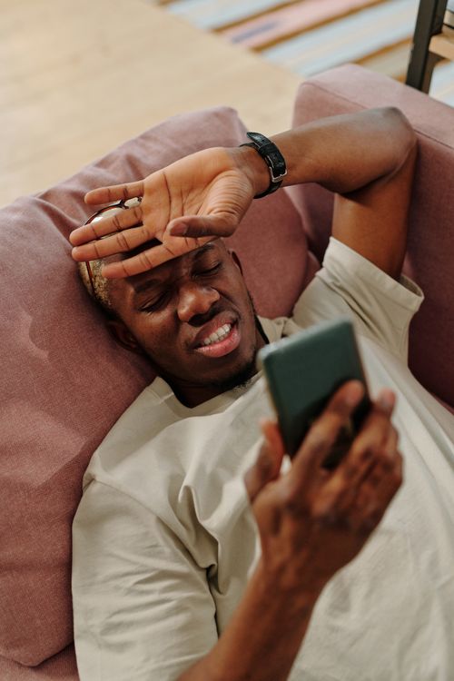 Free An adult black man lies on a sofa, using a smartphone with a relaxed expression indoors. Stock Photo