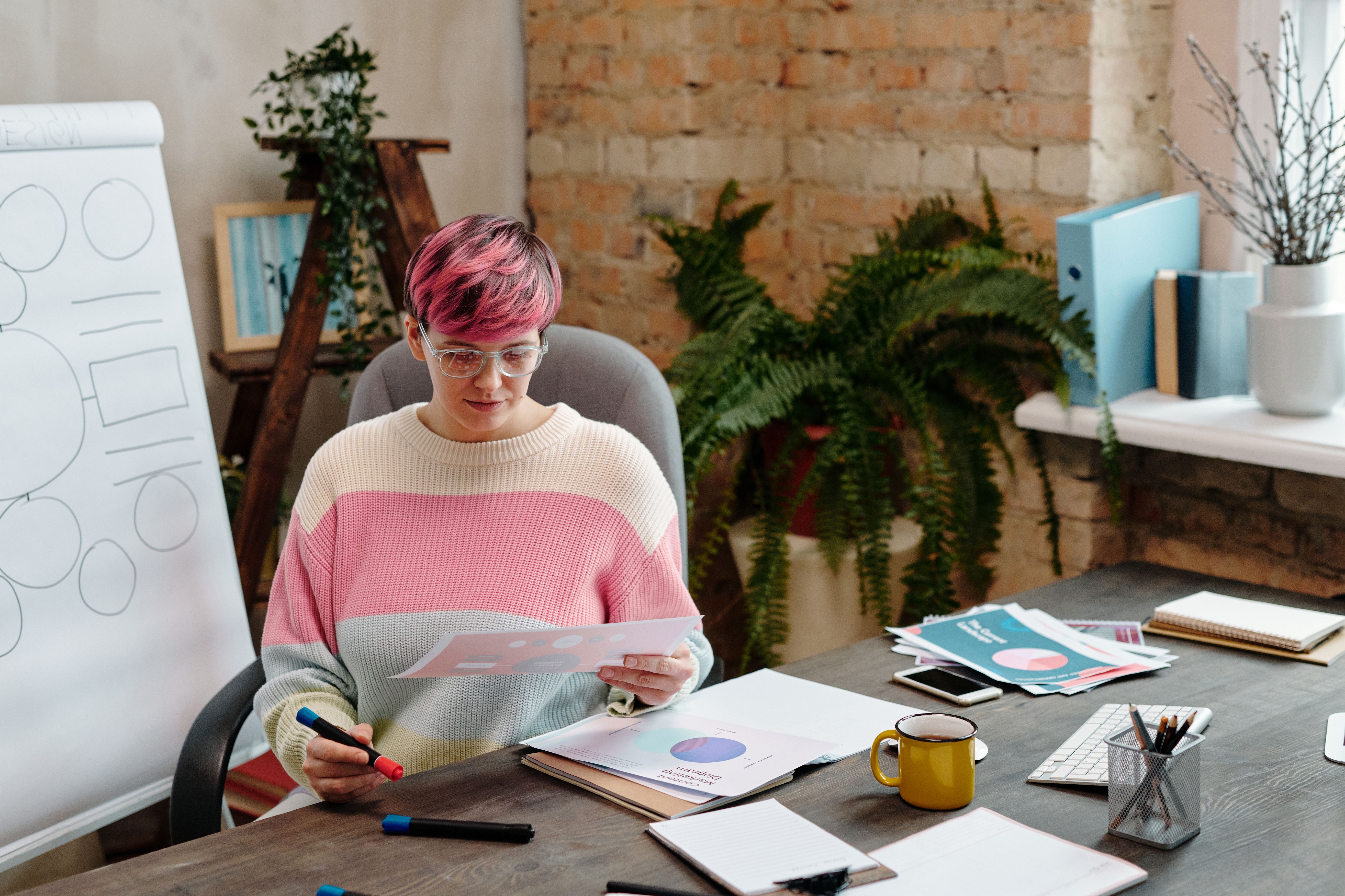A creative professional with pink hair and glasses reviews charts in a modern office setting.
