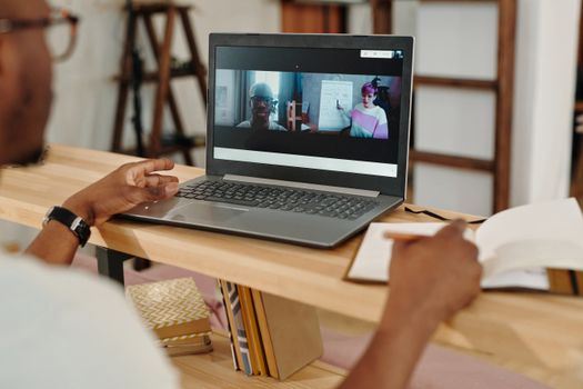 Adult using a laptop for video conferencing, taking notes during a remote meeting indoors.