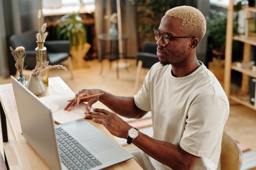 Free Young man working at a desk with a laptop and notebook, writing with pencil. Indoors in a modern office setting. Stock Photo