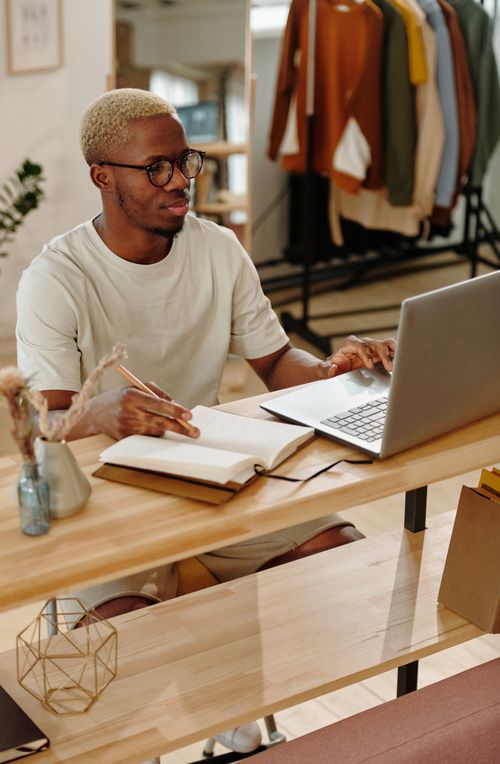 Free A man sitting at a desk writing in a notebook while using a laptop indoors, focused on work. Stock Photo