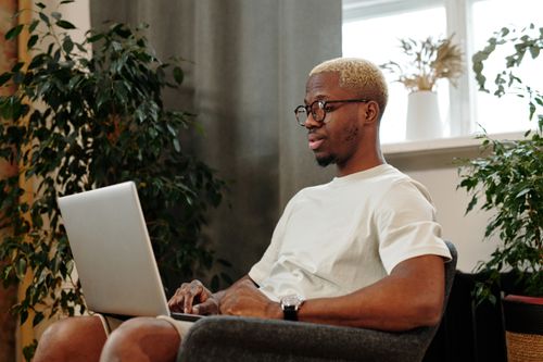 Free Focused young man with glasses working on a laptop in a cozy indoor space surrounded by plants. Stock Photo