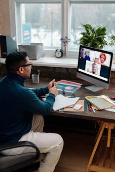 Man sitting at a desk engaged in a video call, communicating with colleagues on a computer screen.