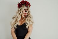 Gorgeous young female with long blond hair in black dress with Mexican Catrina inspired makeup and floral headband standing against white background and looking away