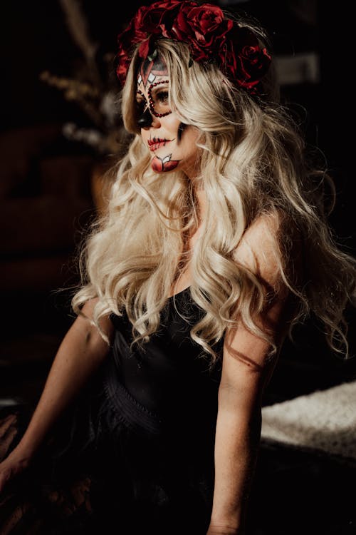 Sensual young lady with long blond hair and sugar skull makeup in elegant dress and floral headband looking away in dark room during Dia de los Muertos party