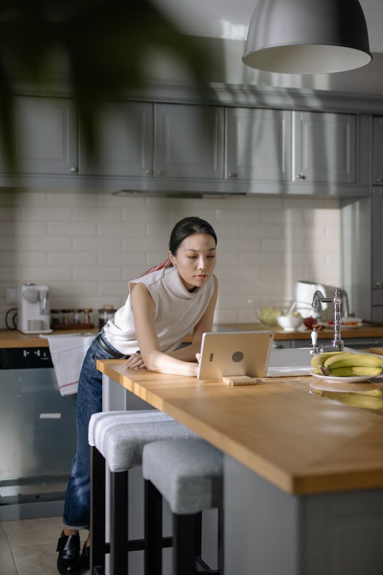 Woman In The Kitchen Looking At Her Tablet While Leaning Against The Table