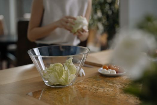 Person preparing fresh salad with lettuce in a glass bowl in a modern kitchen setting.