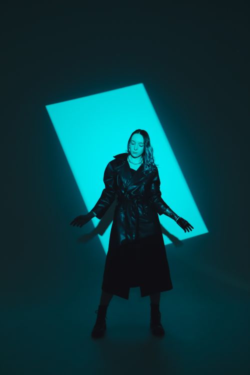 Free Caucasian woman in a black coat posing in a dramatic studio setting with teal lighting. Stock Photo