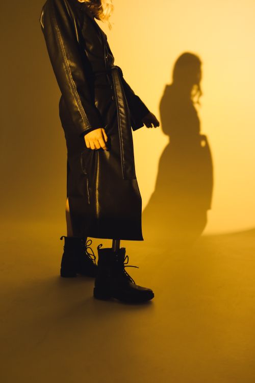 Free Stylish woman in a black leather coat and boots poses against a warm yellow backdrop. Stock Photo