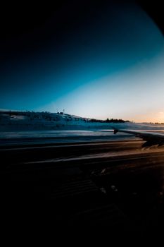 View through porthole on wing of airplane taking off from runway on snowy airfield at cloudless winter sunset