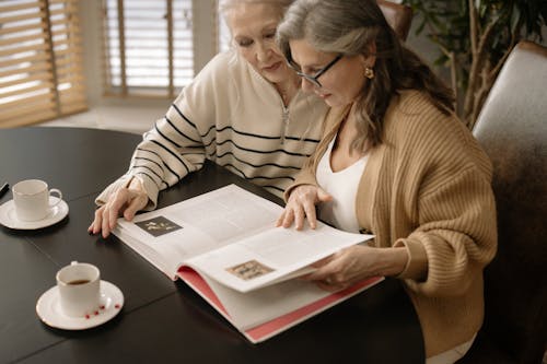 Free Woman in Brown Cardigan Sitting beside Woman in White and Beige Sweater while Reading a Book Stock Photo