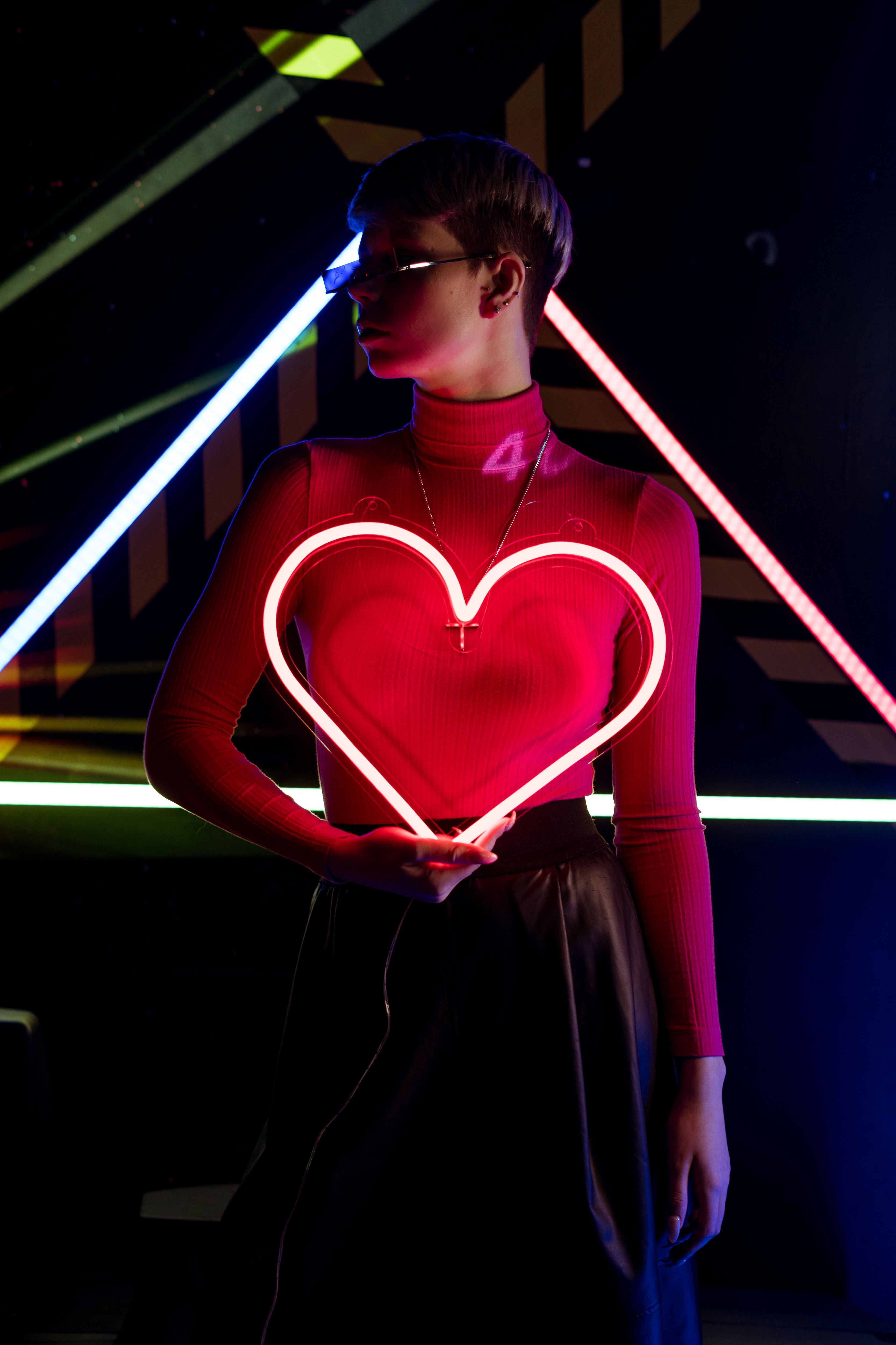 A woman in a turtleneck holds a neon heart light in a cyberpunk setting.