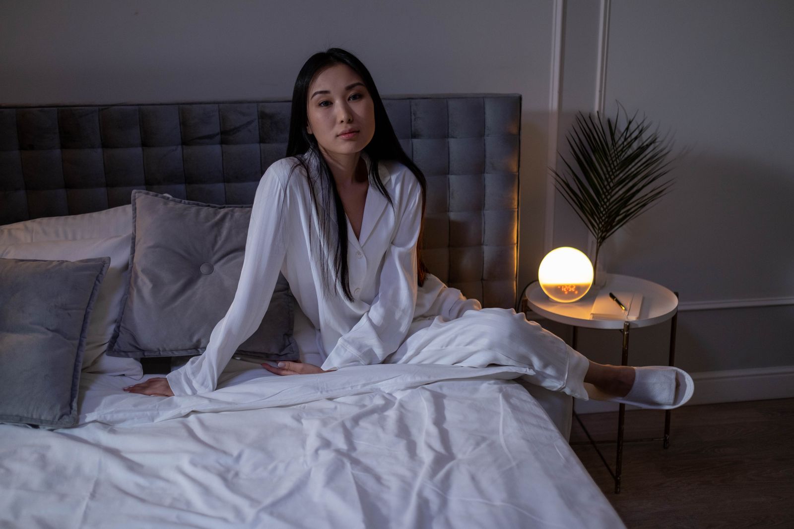 Woman with long dark hair in white satin pajamas sitting on a bed beside a glowing lamp