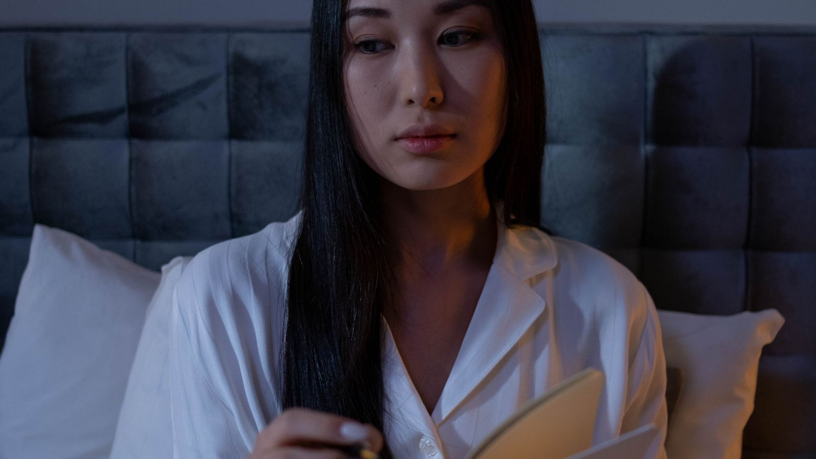 A woman in pajamas writing in a journal while sitting on a bed at night