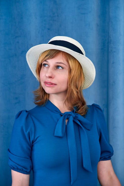 Charming female model with wavy hair wearing blue dress and hat looking away dreamily