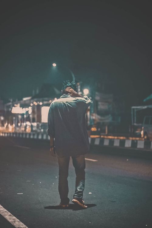 Free A man walks alone on a dimly lit city street at night, with streetlights illuminating the road. Stock Photo