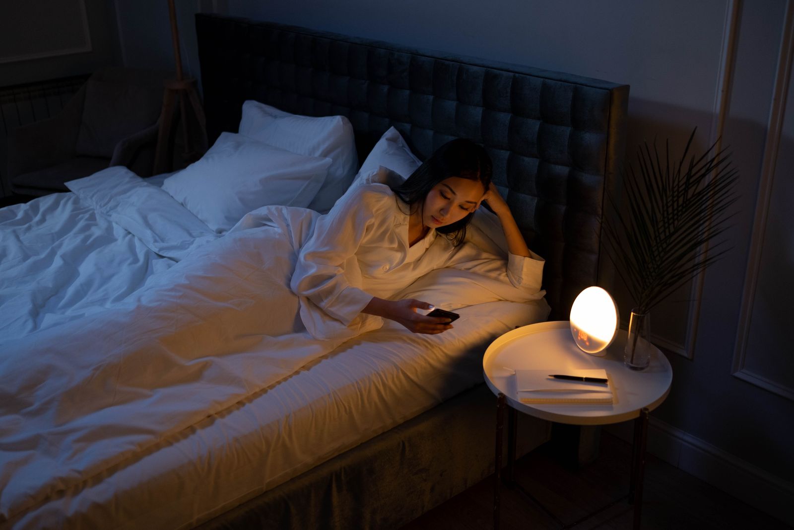 A woman lying in bed at night looking at her phone, lit by a bedside lamp