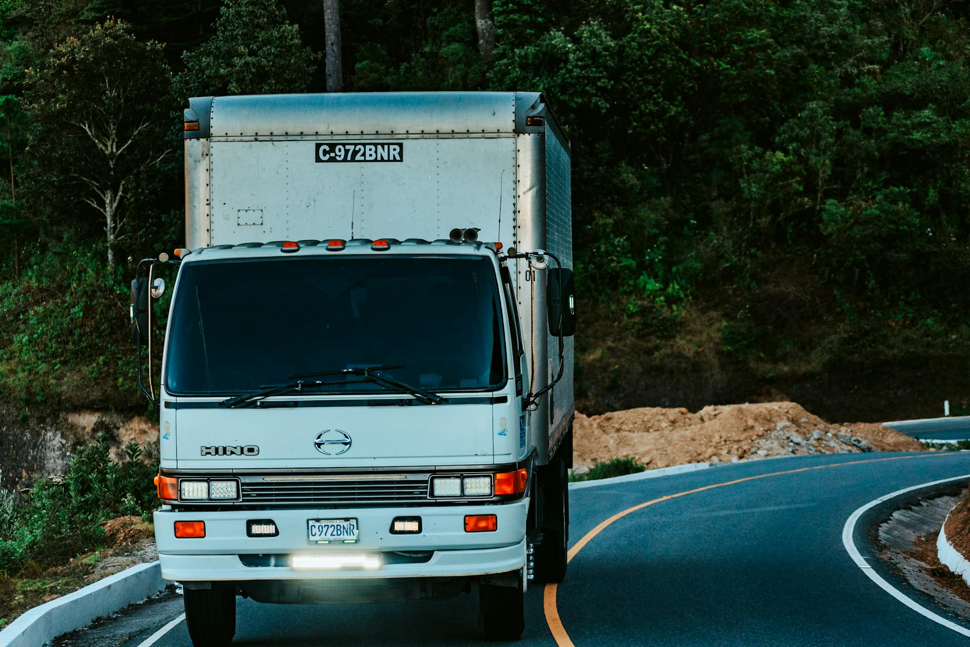 A white truck driving on a winding road surrounded by dense green forest plants.