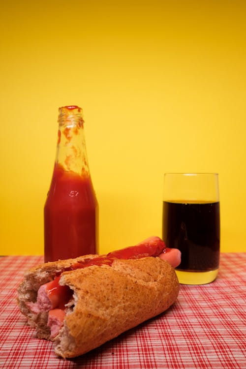 Appetizing homemade hot dog placed on table with bottle of ketchup and glass of coke against yellow background