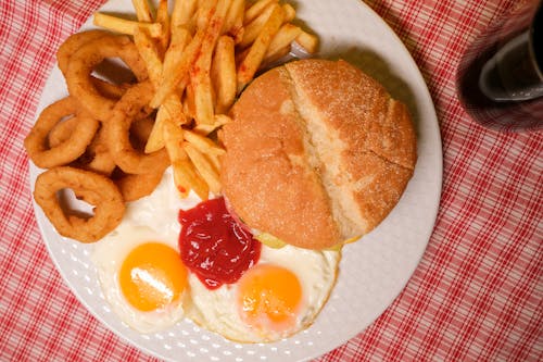 Top view of delicious hamburger with French fires and onion rings served on plate with eggs and ketchup near glass of coke