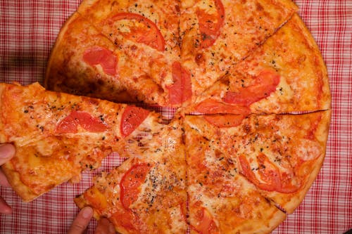 Top view of crop anonymous person taking slice of yummy pizza with tomato and oregano placed on checkered tablecloth