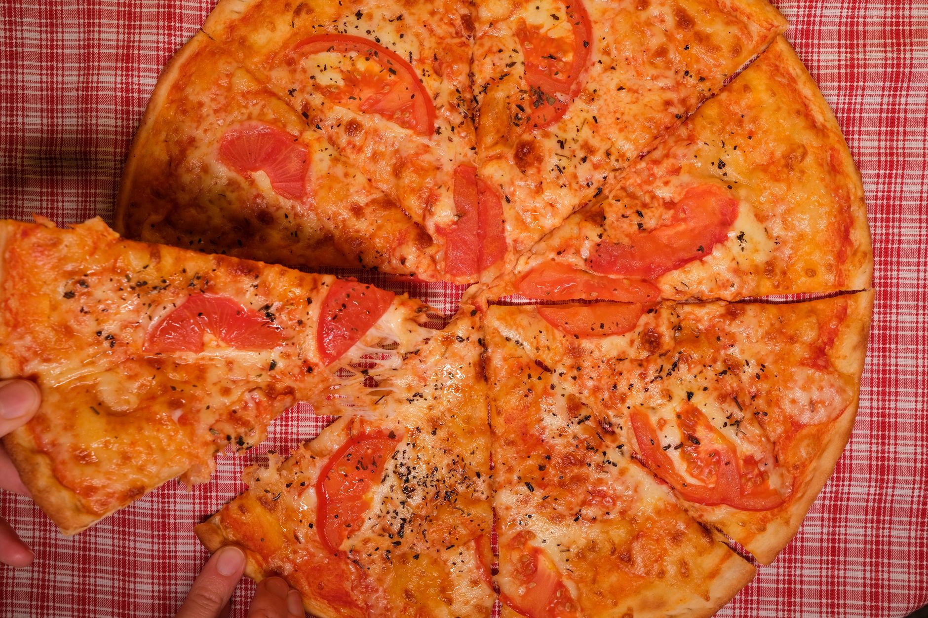 Top view of crop anonymous person taking slice of yummy pizza with tomato and oregano placed on checkered tablecloth