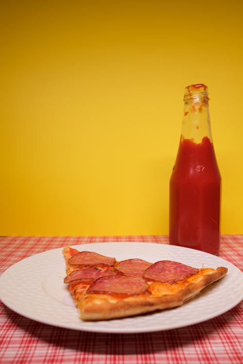 Slice of pizza placed on plate served on table with bottle of ketchup against yellow background