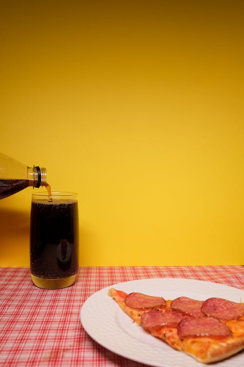 Slice of peperoni pizza on plate near glass with soda on table near yellow wall