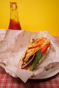 High angle of gyro with french fries and fresh vegetables in parchment paper on table with ketchup bottle against yellow background
