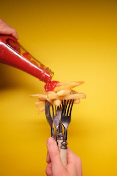 Crop anonymous person with forks in hand pouring ketchup from bottle into fried potatoes on yellow background in light studio