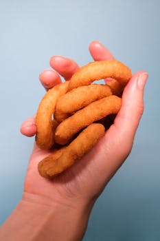 Crop faceless person demonstrating delicious fried onion rings in hand on blue background in light studio
